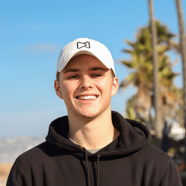 MediaKits Revelry A man smiling, wearing a white baseball cap and a black hoodie, standing in front of palm trees.
