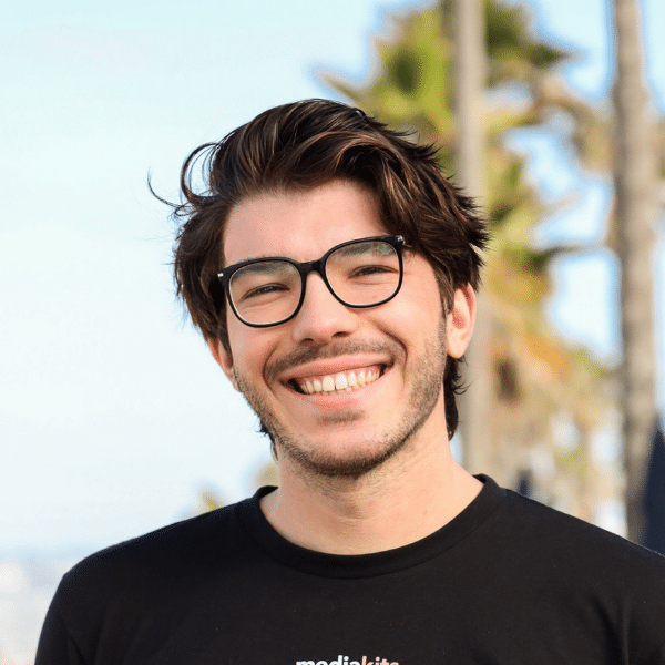 MediaKits Revelry A man with short black hair and glasses, wearing a black T-shirt, smiling and standing in front of palm trees.